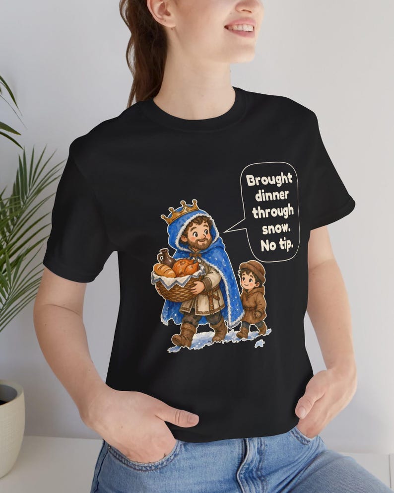 May include: A black t-shirt featuring a graphic of a bearded person wearing a blue crown and cape, carrying a basket of food, walking with a child in the snow. A speech bubble contains the text: Brought dinner through snow. No tip.