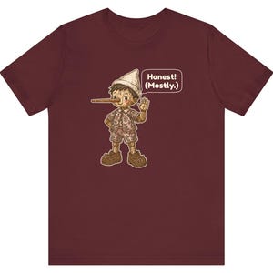 K&ouml;nnte beinhalten: Ein bordeauxrotes Kurzarm-T-Shirt mit einer Grafik einer Holzpuppe mit langer Nase, die einen Hut, ein Hemd und eine kurze Hose tr&auml;gt. Die Figur winkt und eine Sprechblase neben ihr enth&auml;lt den Text, Honest! (Mostly.)