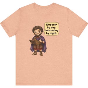 K&ouml;nnte beinhalten: Ein pfirsichfarbenes T-Shirt mit einer Cartoon-Illustration einer b&auml;rtigen Figur in r&ouml;mischer R&uuml;stung und violettem Umhang, die ein Tagebuch h&auml;lt. Eine Sprechblase enth&auml;lt den Text: Emperor by day. Journaling by night.