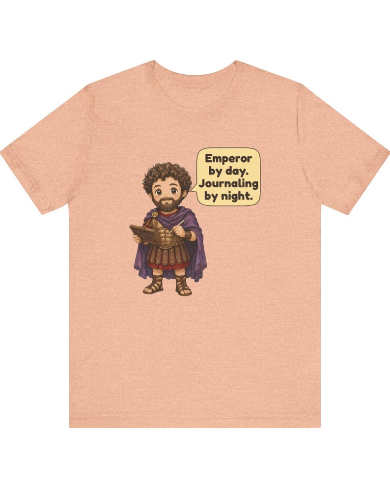 K&ouml;nnte beinhalten: Ein pfirsichfarbenes T-Shirt mit einer Cartoon-Illustration einer b&auml;rtigen Figur in r&ouml;mischer R&uuml;stung und violettem Umhang, die ein Tagebuch h&auml;lt. Eine Sprechblase enth&auml;lt den Text: Emperor by day. Journaling by night.