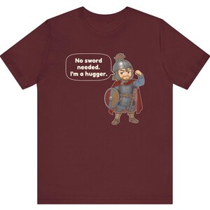 May include: A maroon t-shirt featuring a cartoon illustration of a warrior in chainmail, a helmet, and a red cape, holding a round shield. A small animal sits on the helmet. The text reads: No sword needed. I'm a hugger.