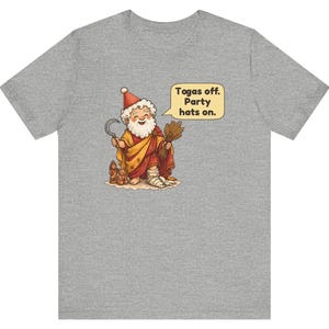 May include: A heather gray t-shirt featuring a cartoon illustration of a bearded figure in a red and yellow robe, holding a sickle and wheat. A speech bubble contains the text: Togas off. Party hats on. The figure wears a red party hat.