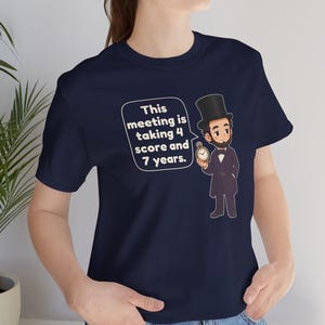 May include: A navy blue t-shirt featuring a cartoon illustration of Abraham Lincoln wearing a top hat and holding a pocket watch. A speech bubble next to the figure contains the text: This meeting is taking 4 score and 7 years.