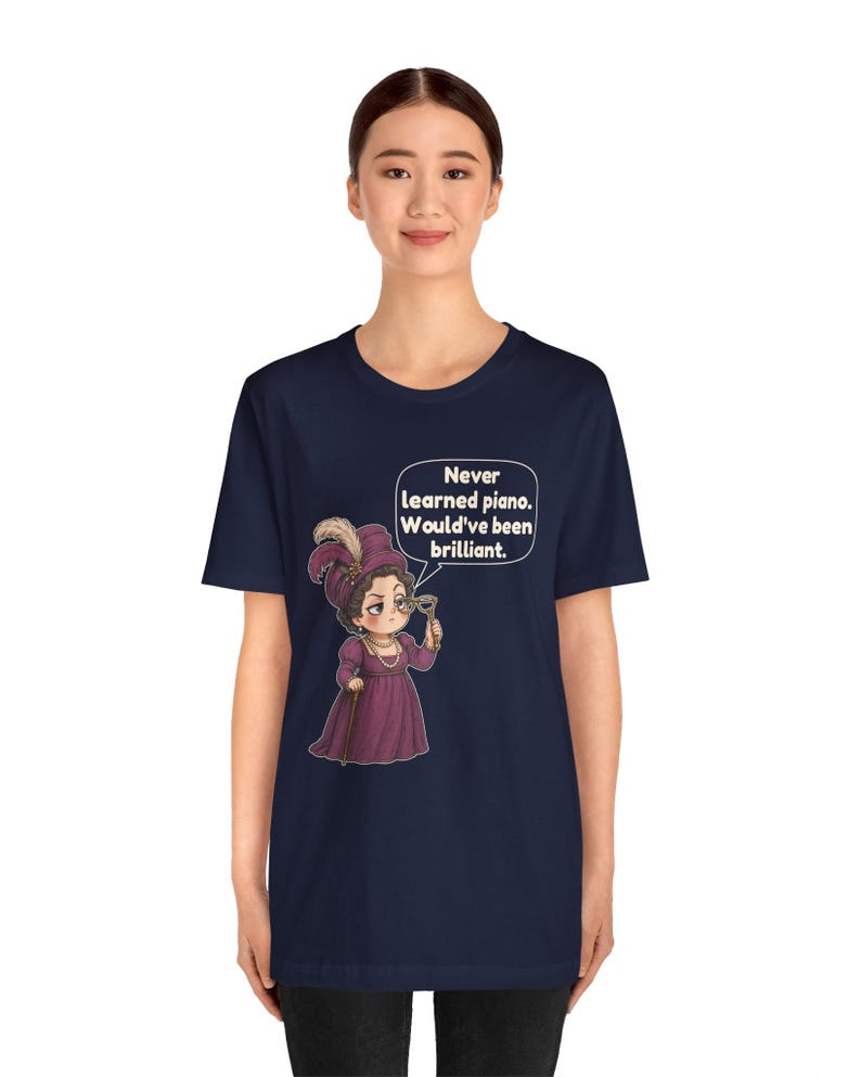 May include: A navy blue short-sleeve t-shirt featuring a cartoon illustration of a person in a purple dress and hat holding a monocle. A speech bubble contains the text: Never learned piano. Would've been brilliant.