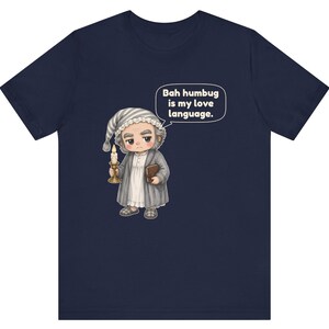 Puede incluir: Una camiseta azul marino de manga corta con una ilustraci&oacute;n de dibujos animados de un personaje con gorro de dormir y bata que sostiene una vela y un libro. Un globo de texto sobre el personaje contiene el texto, Bah humbug is my love language.