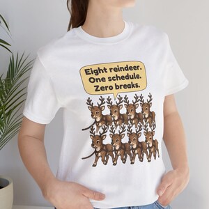 K&ouml;nnte beinhalten: Ein wei&szlig;es Kurzarm-T-Shirt mit einer Grafik von acht braunen Rentieren, die in zwei Reihen angeordnet sind. &Uuml;ber den Rentieren befindet sich eine gelbe Sprechblase mit dem schwarzen Text: Eight reindeer. One schedule. Zero breaks.