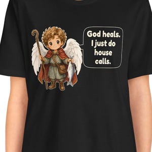 May include: A black t-shirt featuring a graphic of a curly-haired angel with white wings, wearing a red cloak and brown tunic, holding a wooden staff and a fish. A speech bubble next to the angel contains the text: God heals. I just do house calls.
