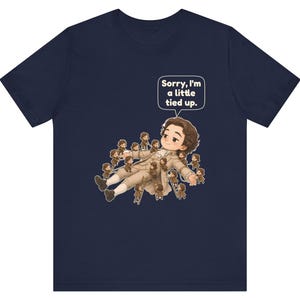 Op de afbeelding: Een marineblauw t-shirt met een afbeelding van een groot personage dat ligt, omringd door vele kleine figuurtjes die ladders gebruiken om het personage vast te binden. Een tekstballon bevat de tekst, Sorry, I'm a little tied up.