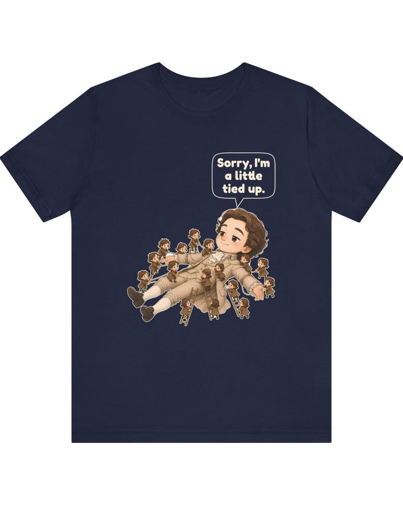 Op de afbeelding: Een marineblauw t-shirt met een afbeelding van een groot personage dat ligt, omringd door vele kleine figuurtjes die ladders gebruiken om het personage vast te binden. Een tekstballon bevat de tekst, Sorry, I'm a little tied up.