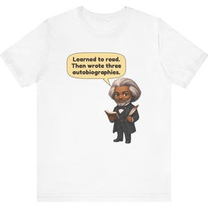 Puede incluir: Una camiseta blanca con una ilustraci&oacute;n de dibujos animados de Frederick Douglass sosteniendo un libro y una pluma. Un globo de texto sobre la figura contiene el texto: Learned to read. Then wrote three autobiographies.