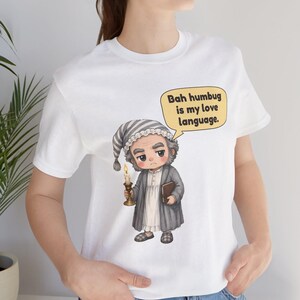 Puede incluir: Una camiseta blanca con el gr&aacute;fico de un personaje de dibujos animados con gorro de dormir y bata a rayas, sosteniendo una vela encendida y un libro. Un bocadillo amarillo contiene el texto negro, Bah humbug is my love language.
