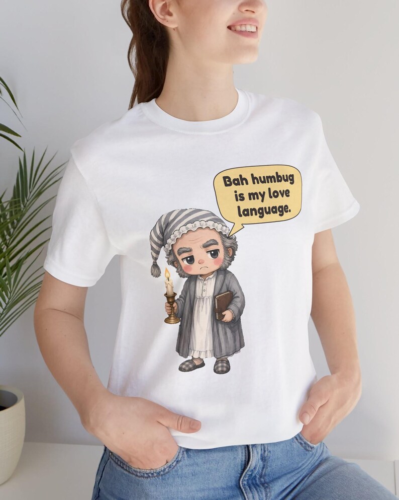 Puede incluir: Una camiseta blanca con el gr&aacute;fico de un personaje de dibujos animados con gorro de dormir y bata a rayas, sosteniendo una vela encendida y un libro. Un bocadillo amarillo contiene el texto negro, Bah humbug is my love language.