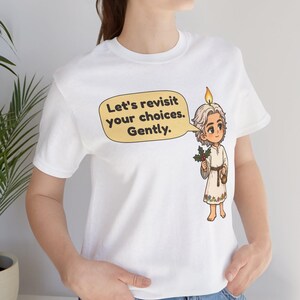 May include: A white short-sleeved t-shirt featuring a cartoon illustration of a character with a candle flame on their head, holding a sprig of holly. A yellow speech bubble contains the text: Let's revisit your choices. Gently.