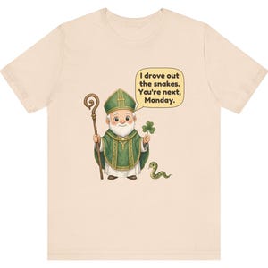 Puede incluir: Una camiseta de color beige claro con una ilustraci&oacute;n de dibujos animados de una figura con vestimentas religiosas verdes que sostiene un bast&oacute;n de madera y un tr&eacute;bol. Una peque&ntilde;a serpiente verde est&aacute; cerca. Un bocadillo contiene el texto: I drove out the snakes. You're next, Monday.
