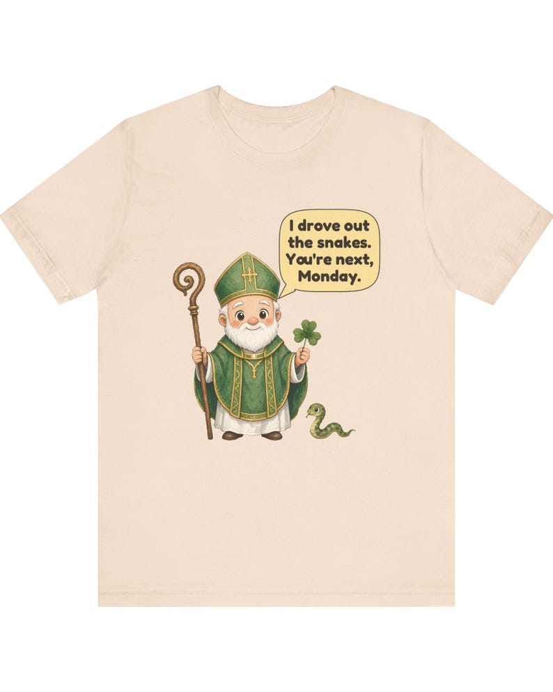 Puede incluir: Una camiseta de color beige claro con una ilustraci&oacute;n de dibujos animados de una figura con vestimentas religiosas verdes que sostiene un bast&oacute;n de madera y un tr&eacute;bol. Una peque&ntilde;a serpiente verde est&aacute; cerca. Un bocadillo contiene el texto: I drove out the snakes. You're next, Monday.