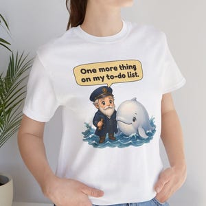 May include: A white t-shirt featuring a graphic of a cartoon sea captain with a white beard and a blue uniform standing in blue ocean waves next to a white whale. Above them is a yellow speech bubble with the text, One more thing on my to-do list.