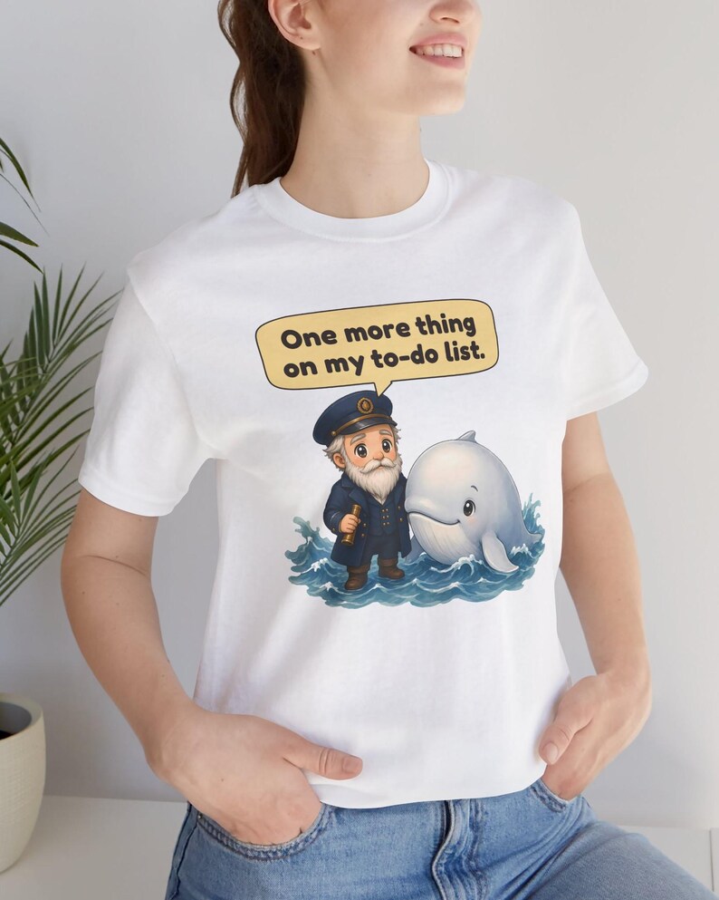 May include: A white t-shirt featuring a graphic of a cartoon sea captain with a white beard and a blue uniform standing in blue ocean waves next to a white whale. Above them is a yellow speech bubble with the text, One more thing on my to-do list.