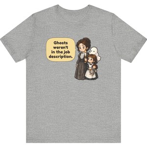 Op de afbeelding: Een grijs gem&ecirc;leerd t-shirt met een cartoonillustratie van een persoon in een donkere jurk die een kaars vasthoudt en een kind met een pop, met een klein wit spookje erachter. Een tekstballon bevat de tekst: Ghosts weren't in the job description.