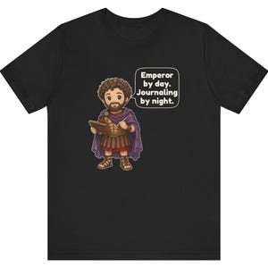 K&ouml;nnte beinhalten: Ein schwarzes T-Shirt mit einer Cartoon-Illustration eines b&auml;rtigen Kaisers in r&ouml;mischer R&uuml;stung und violettem Umhang, der ein Buch und einen Stift h&auml;lt. Eine Sprechblase enth&auml;lt den Text: Emperor by day. Journaling by night.
