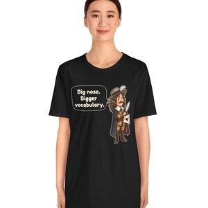 May include: A black short-sleeve t-shirt featuring a graphic of a historical figure with a large nose, wearing a hat and cape, holding a quill and paper. A speech bubble contains the text: Big nose. Bigger vocabulary.