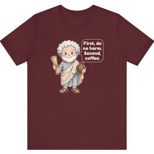 May include: A maroon t-shirt featuring a cartoon illustration of a white-haired, bearded figure in a light blue tunic and beige toga. The figure holds a scroll and a brown pot. A speech bubble contains the text: First, do no harm. Second, coffee.