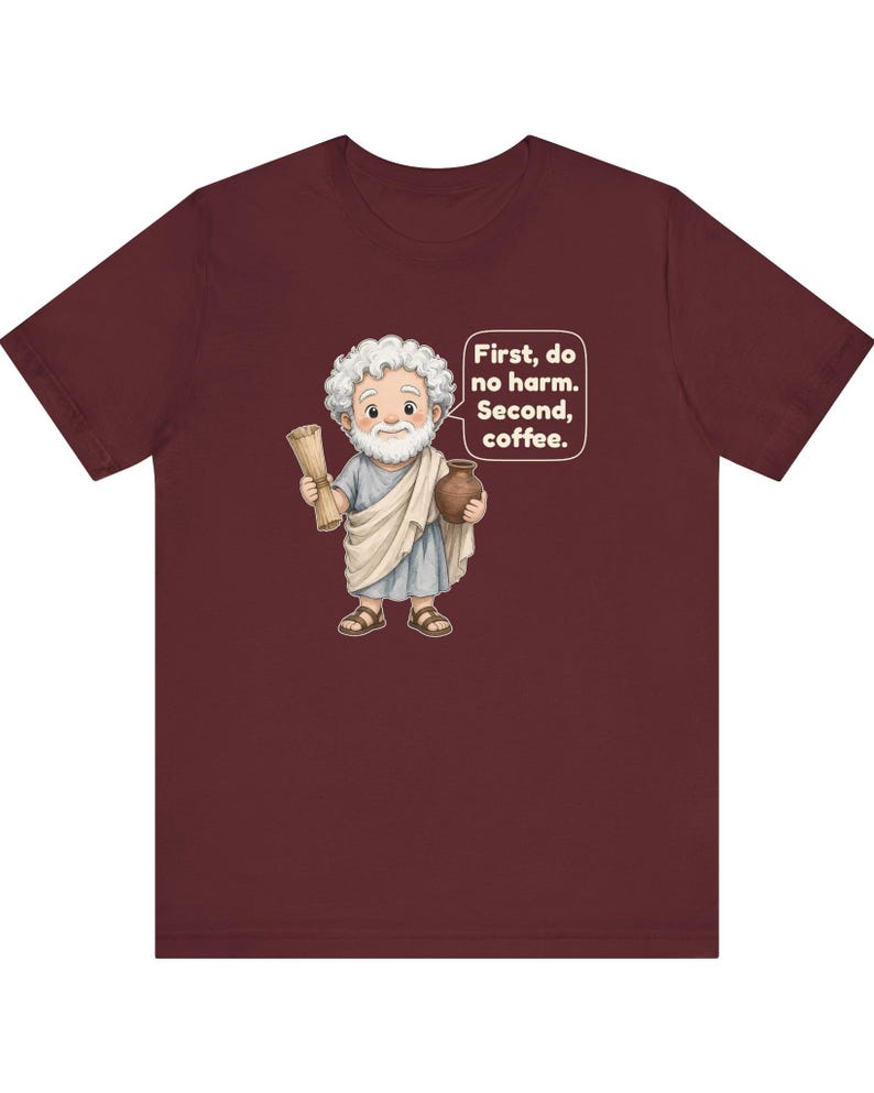 May include: A maroon t-shirt featuring a cartoon illustration of a white-haired, bearded figure in a light blue tunic and beige toga. The figure holds a scroll and a brown pot. A speech bubble contains the text: First, do no harm. Second, coffee.