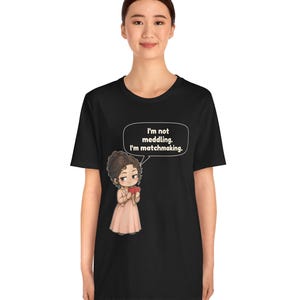 K&ouml;nnte beinhalten: Ein schwarzes T-Shirt mit kurzen &Auml;rmeln und einer Cartoon-Illustration einer Person mit braunen Haaren in einem hellrosa Kleid, die ein rotes Herz h&auml;lt. Eine Sprechblase enth&auml;lt den Text: I'm not meddling. I'm matchmaking.