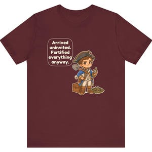 May include: A maroon t-shirt featuring a cartoon illustration of a historical explorer in a blue coat and tricorn hat holding a shovel and compass, with a wooden chest and a speech bubble that reads: Arrived uninvited. Fortified everything anyway.
