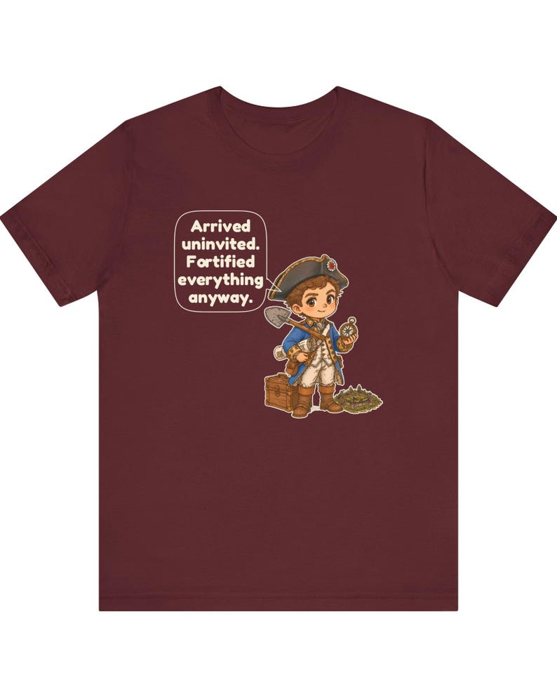 May include: A maroon t-shirt featuring a cartoon illustration of a historical explorer in a blue coat and tricorn hat holding a shovel and compass, with a wooden chest and a speech bubble that reads: Arrived uninvited. Fortified everything anyway.