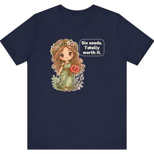 May include: A navy blue t-shirt featuring a cartoon illustration of a character with long brown hair, a flower crown, and a green dress holding a pomegranate. A speech bubble next to the character contains the text: Six seeds. Totally worth it.