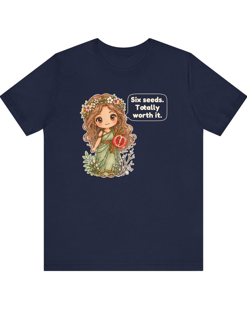 May include: A navy blue t-shirt featuring a cartoon illustration of a character with long brown hair, a flower crown, and a green dress holding a pomegranate. A speech bubble next to the character contains the text: Six seeds. Totally worth it.