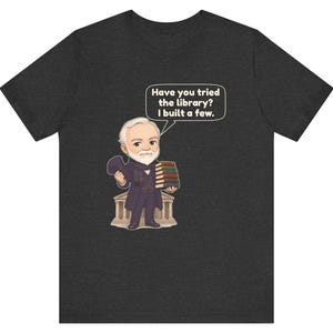 May include: A dark heather gray t-shirt featuring a cartoon illustration of a bearded man in a suit holding a stack of books and a hat. A speech bubble above him contains the text: Have you tried the library? I built a few.