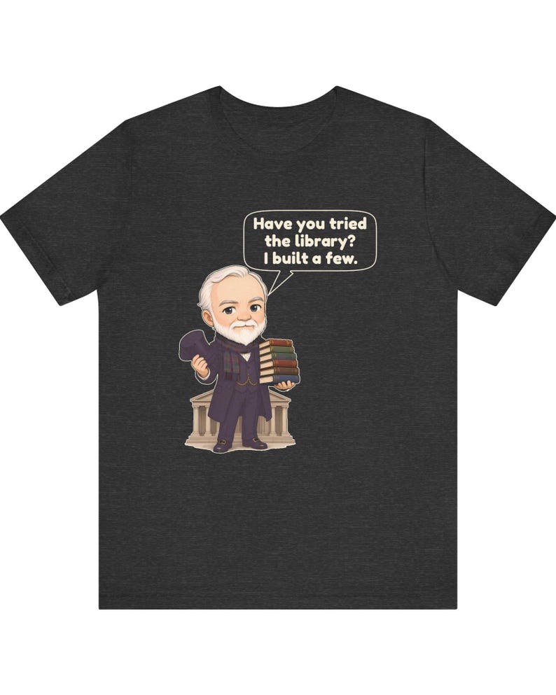 May include: A dark heather gray t-shirt featuring a cartoon illustration of a bearded man in a suit holding a stack of books and a hat. A speech bubble above him contains the text: Have you tried the library? I built a few.