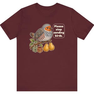 K&ouml;nnte beinhalten: Ein kastanienbraunes T-Shirt mit einer Grafik eines grauen und orangefarbenen Vogels, der auf einem Ast mit zwei Birnen und zwei kleinen Geschenken sitzt. Eine Sprechblase neben dem Vogel enth&auml;lt den Text: "Please stop sending birds."