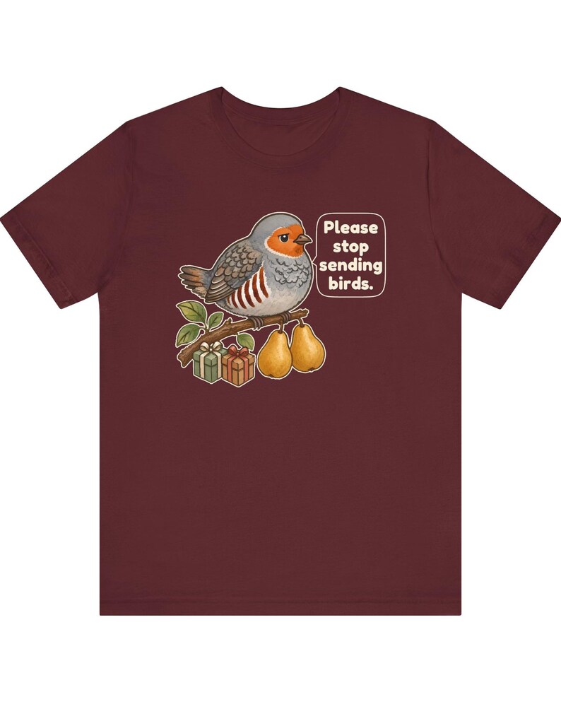K&ouml;nnte beinhalten: Ein kastanienbraunes T-Shirt mit einer Grafik eines grauen und orangefarbenen Vogels, der auf einem Ast mit zwei Birnen und zwei kleinen Geschenken sitzt. Eine Sprechblase neben dem Vogel enth&auml;lt den Text: "Please stop sending birds."