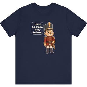 Pu&ograve; includere: Una t-shirt blu navy con la grafica di un soldatino schiaccianoci in uniforme rossa con dettagli dorati. Un fumetto accanto al personaggio riporta il testo: Hard to crack. Easy to love. Il disegno &egrave; centrato sulla parte anteriore.