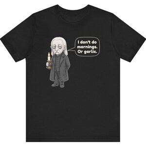 May include: A black short-sleeved t-shirt featuring a graphic of a pale, tired-looking character in a dark coat holding a lit candle. A speech bubble next to the character contains the text: I don't do mornings. Or garlic.