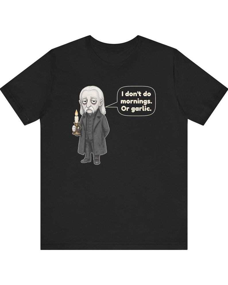 May include: A black short-sleeved t-shirt featuring a graphic of a pale, tired-looking character in a dark coat holding a lit candle. A speech bubble next to the character contains the text: I don't do mornings. Or garlic.