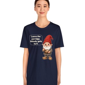 May include: A navy blue short-sleeve t-shirt featuring a graphic of a gnome wearing a red hat and holding a bowl of porridge. A speech bubble next to the gnome contains the text: Leave the porridge. Nobody gets hurt.