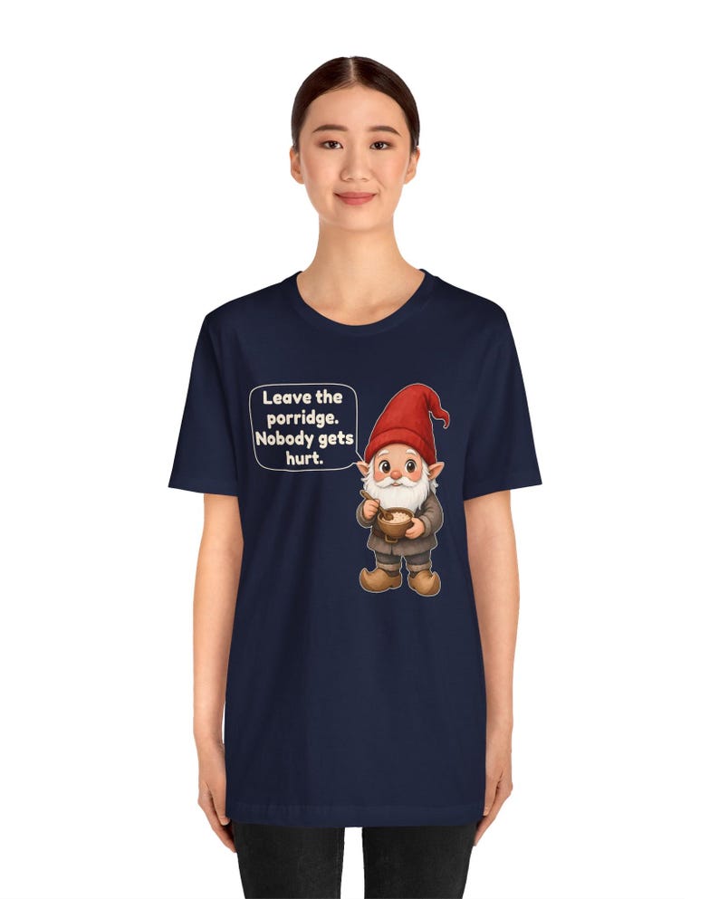 May include: A navy blue short-sleeve t-shirt featuring a graphic of a gnome wearing a red hat and holding a bowl of porridge. A speech bubble next to the gnome contains the text: Leave the porridge. Nobody gets hurt.