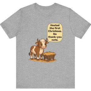 以下が含まれることがあります： ヘザーグレーのTシャツ。干し草が入った木製の飼い葉桶の横に立つ、茶色と白の子牛の漫画が描かれています。子牛の上の吹き出しには「Hosted the first Christmas. No thank-you note.」というテキストがあります。
