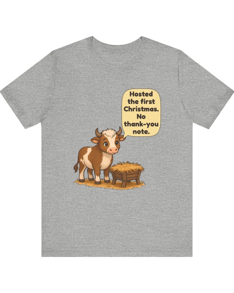 以下が含まれることがあります： ヘザーグレーのTシャツ。干し草が入った木製の飼い葉桶の横に立つ、茶色と白の子牛の漫画が描かれています。子牛の上の吹き出しには「Hosted the first Christmas. No thank-you note.」というテキストがあります。