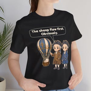 May include: A black t-shirt featuring a graphic of a blue and white hot air balloon carrying a sheep, accompanied by two figures in 18th-century attire. A speech bubble above contains the text: The sheep flew first. Obviously.
