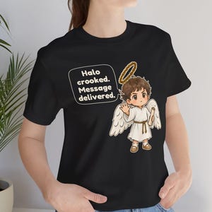 May include: A black t-shirt featuring a graphic of a cartoon angel with brown hair, white wings, and a halo. The angel holds a scroll and has a speech bubble that reads: Halo crooked. Message delivered. The design is centered on the chest area.