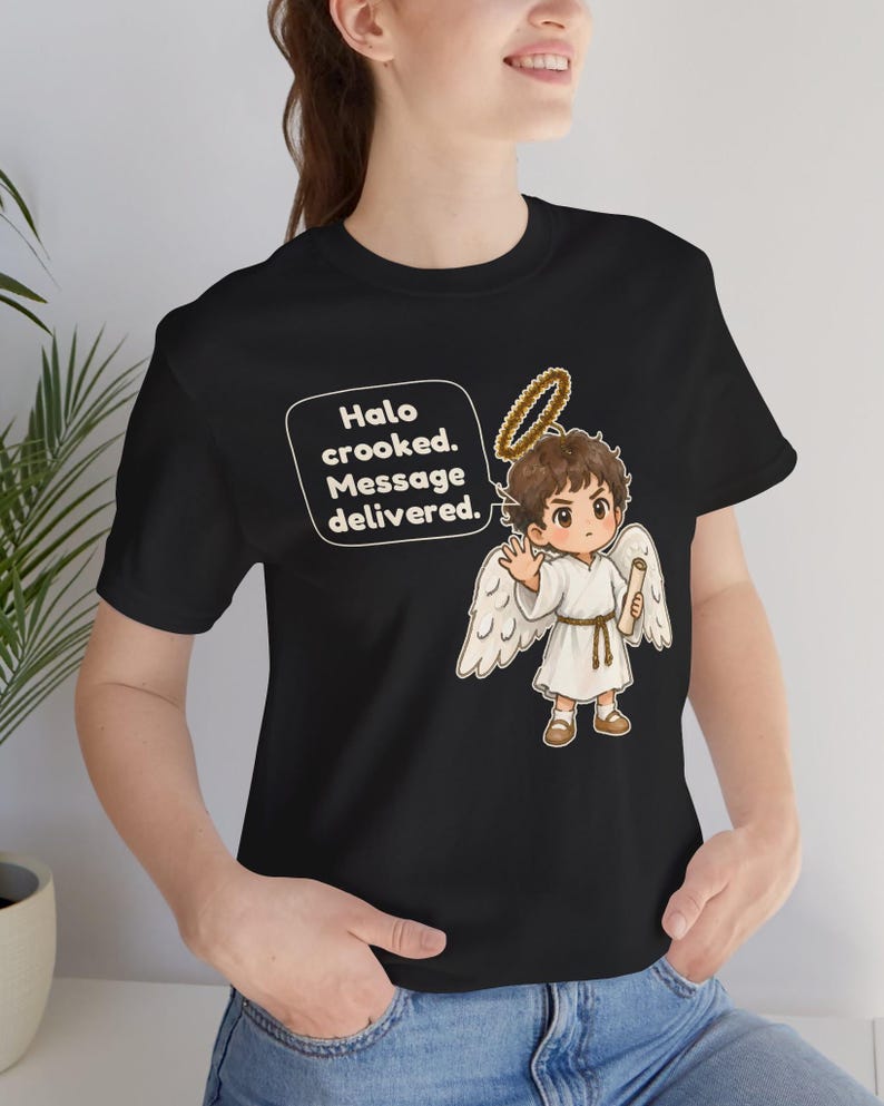 May include: A black t-shirt featuring a graphic of a cartoon angel with brown hair, white wings, and a halo. The angel holds a scroll and has a speech bubble that reads: Halo crooked. Message delivered. The design is centered on the chest area.