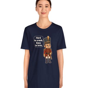Pu&ograve; includere: Una t-shirt blu navy a maniche corte con la grafica di un soldato schiaccianoci in uniforme rossa. Accanto al soldato c'&egrave; un fumetto con il testo: Hard to crack. Easy to love. Il disegno &egrave; centrato sulla parte del petto.