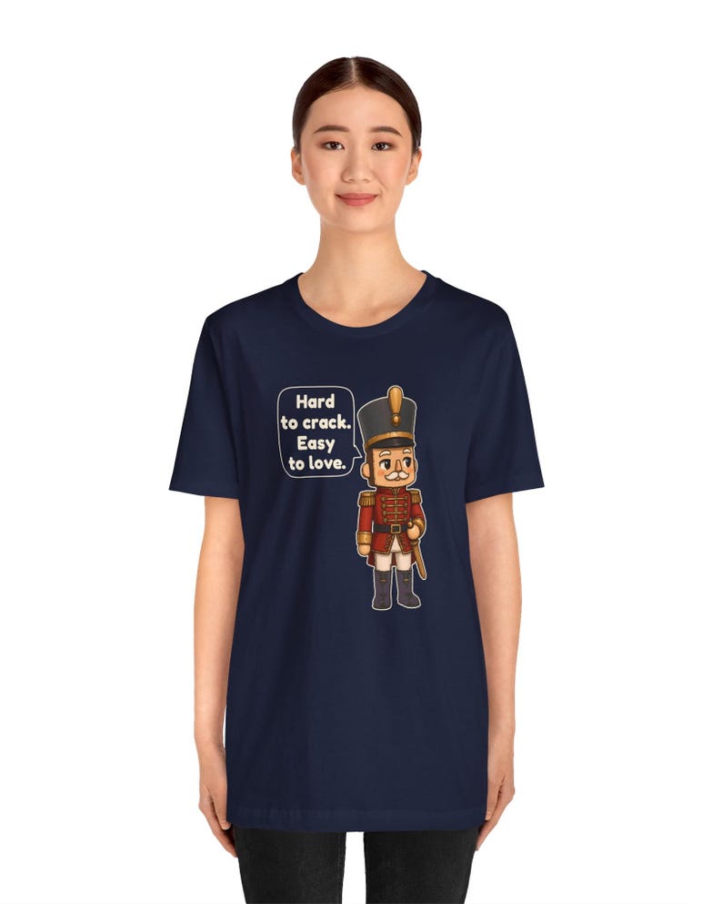 Pu&ograve; includere: Una t-shirt blu navy a maniche corte con la grafica di un soldato schiaccianoci in uniforme rossa. Accanto al soldato c'&egrave; un fumetto con il testo: Hard to crack. Easy to love. Il disegno &egrave; centrato sulla parte del petto.