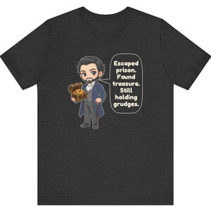 May include: A dark heather gray t-shirt featuring a cartoon illustration of a bearded man in a blue coat holding a treasure chest, with a speech bubble containing the text: Escaped prison. Found treasure. Still holding grudges.