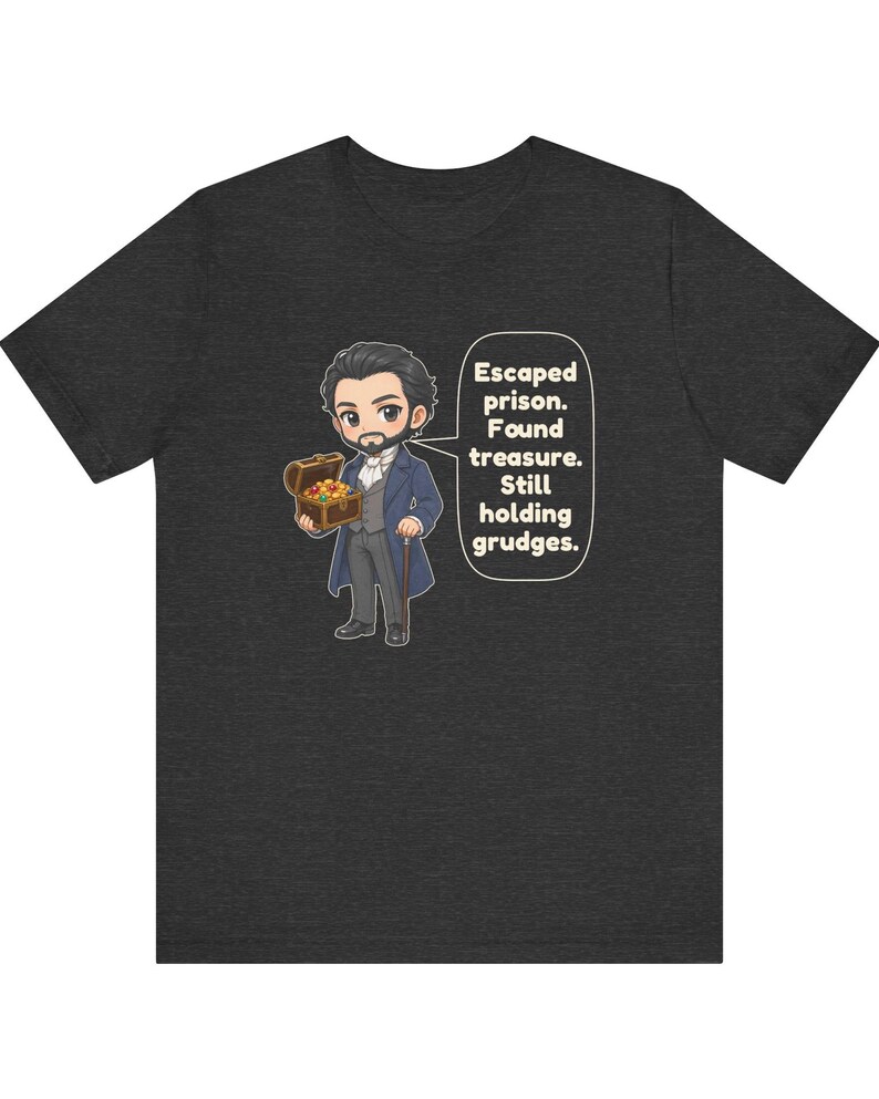 May include: A dark heather gray t-shirt featuring a cartoon illustration of a bearded man in a blue coat holding a treasure chest, with a speech bubble containing the text: Escaped prison. Found treasure. Still holding grudges.