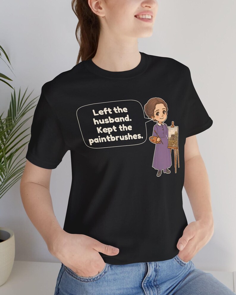 May include: A black t-shirt featuring a cartoon illustration of a person in a purple dress standing next to an easel with a painting. A speech bubble contains the text: Left the husband. Kept the paintbrushes.